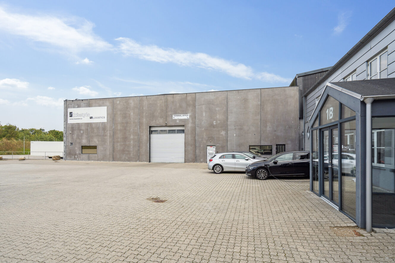 En moderne industribygning med en grå facade, en stor garageport, to parkerede biler og en klar himmel over.