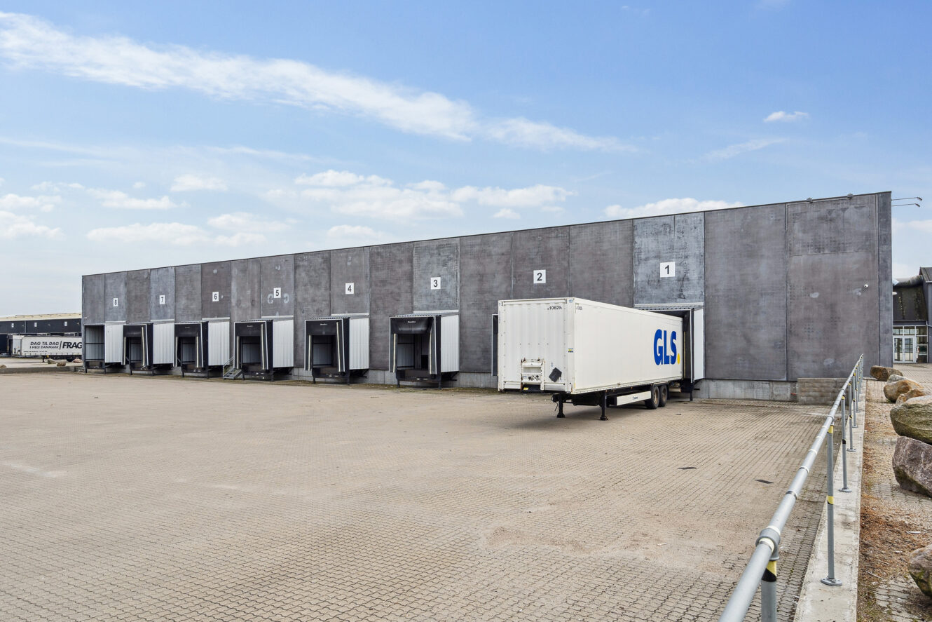 En hvid GLS-lastvognstrailer er parkeret ved en af flere læsseramper på siden af et gråt industrilager under en delvis overskyet himmel.