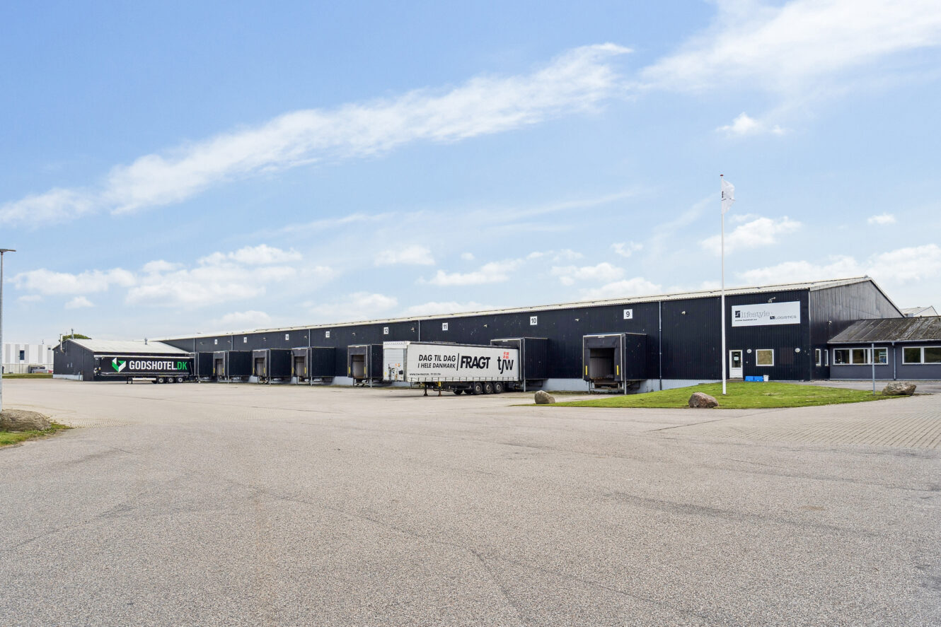 En stor, mørk lagerbygning med flere læsseramper og tre lastbiler parkeret udenfor på en klar dag.