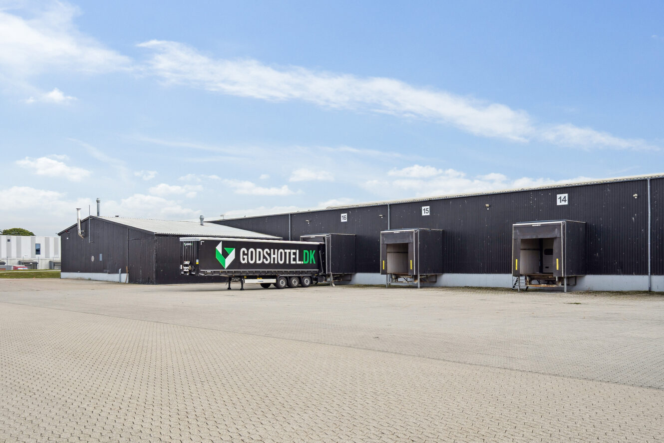En sort lagerbygning med tre læsseramper og en GODSHOTEL.DK-trailer parkeret udenfor på en asfalteret plads under en delvis overskyet himmel.