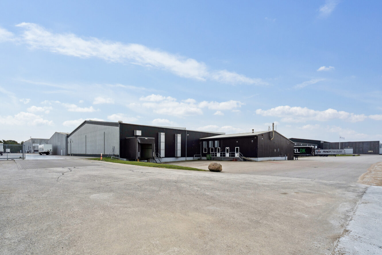 Et stort industrielt lagerkompleks med grå bygninger, et bredt asfalteret område og minimal beplantning under en delvis overskyet himmel.