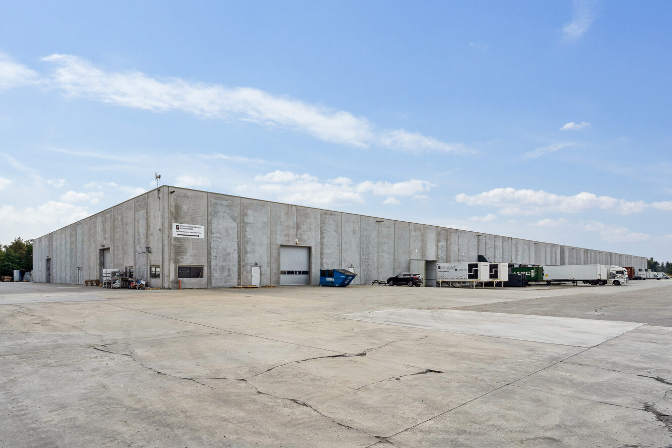Stort industrilager med betonfacade, flere læsseramper, kommercielle lastbiler og containere på en stor asfalteret grund under en delvis overskyet himmel.