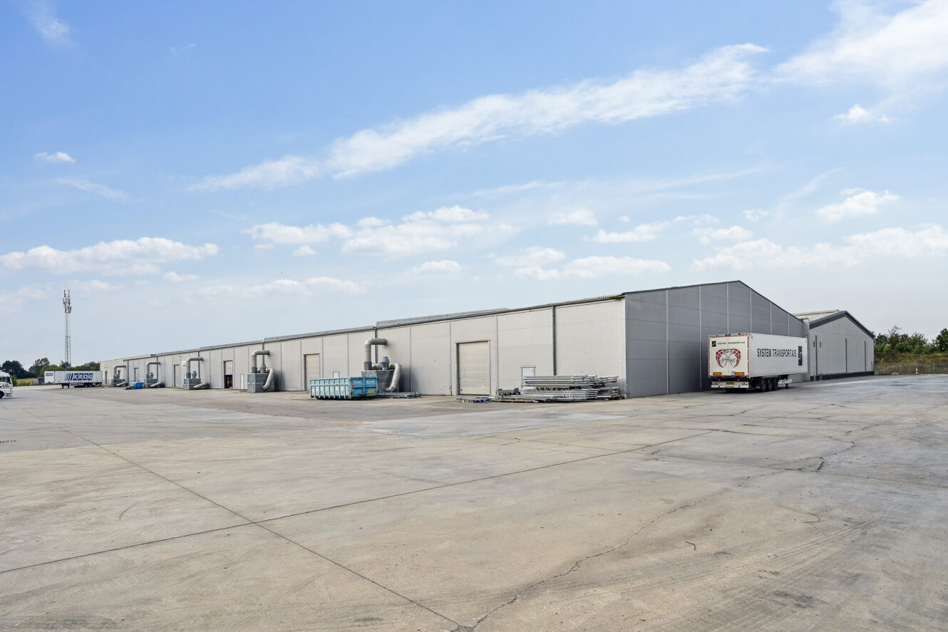 Store lagerbygninger med læsseramper og en parkeret lastbiltrailer på en stor, tom betonplads under en delvis overskyet himmel.