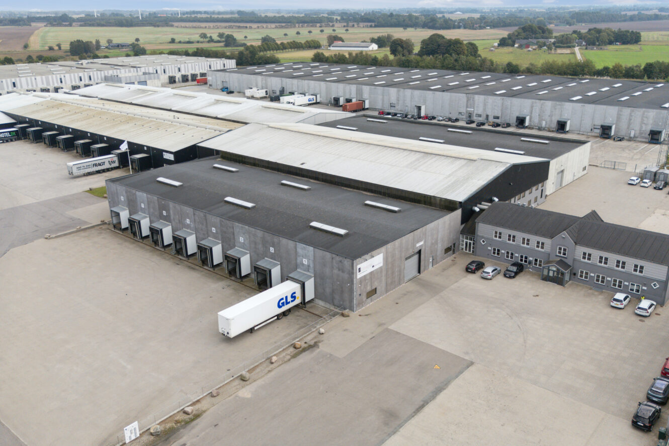 Luftfoto af et stort industrielt lagerkompleks med flere læsseramper, parkerede lastbiler og tilstødende kontorbygninger, omgivet af åbent land og marker.