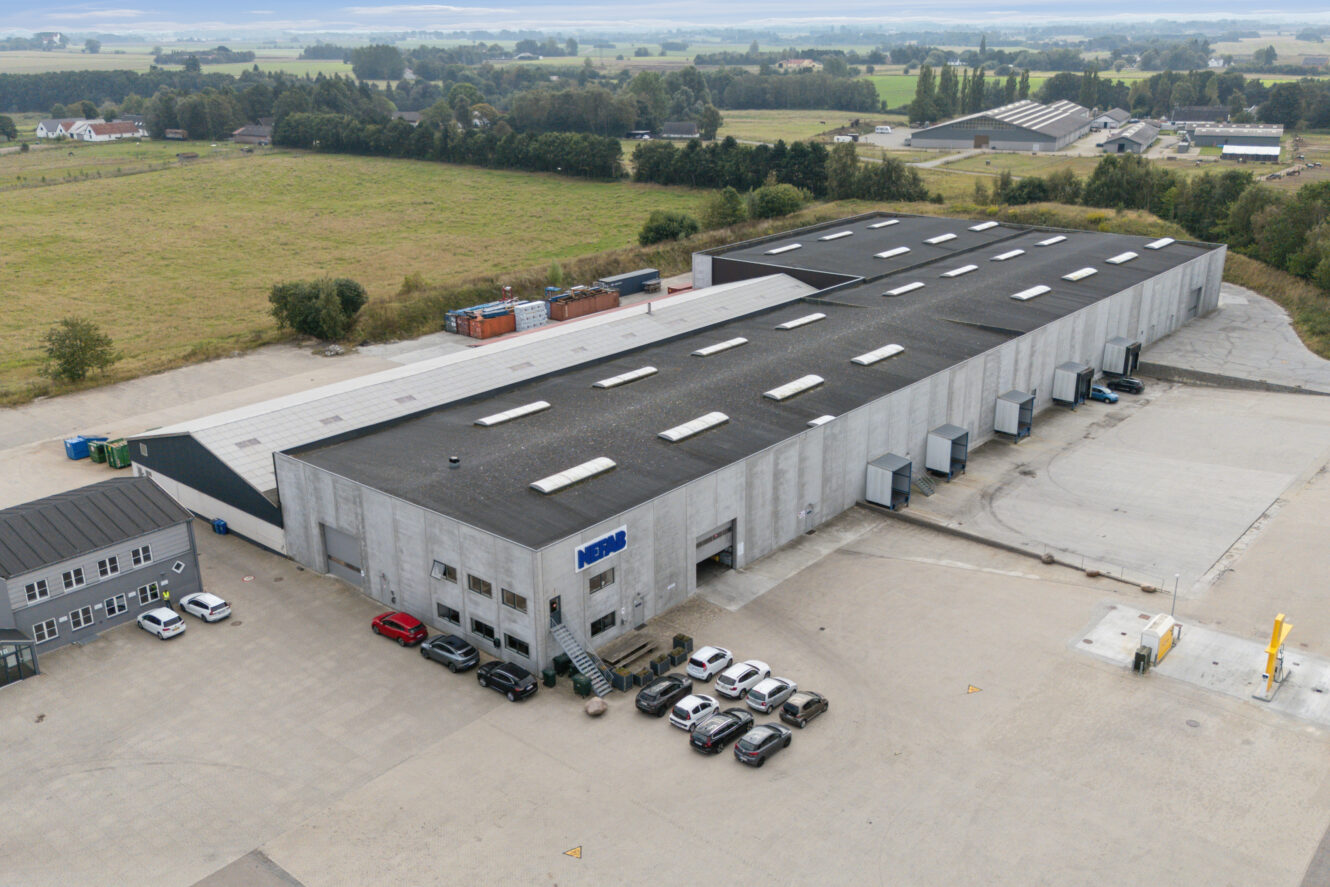 Luftfoto af et stort industrilager med flere læsseramper, parkerede biler foran og åbne marker omkring bygningen.