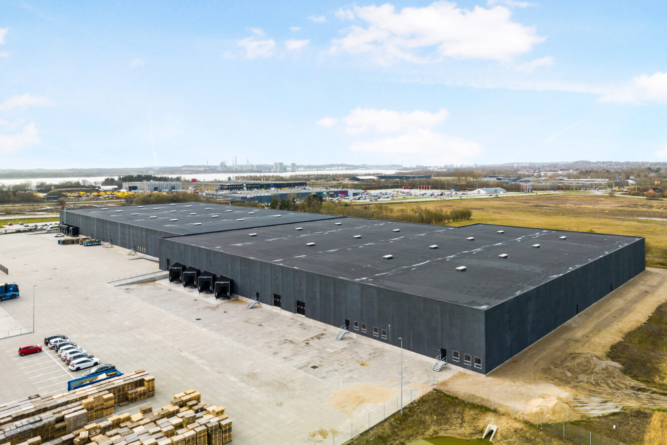 Luftfoto af en stor sort lagerbygning med flere læsseramper, omgivet af parkerede biler, paller og åbent land under en blå himmel.