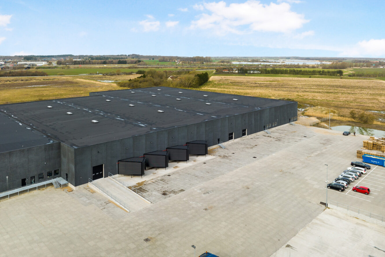 En stor sort lagerbygning med flere læsseramper og en parkeringsplads i et landligt område med marker og nogle få parkerede biler i nærheden.
