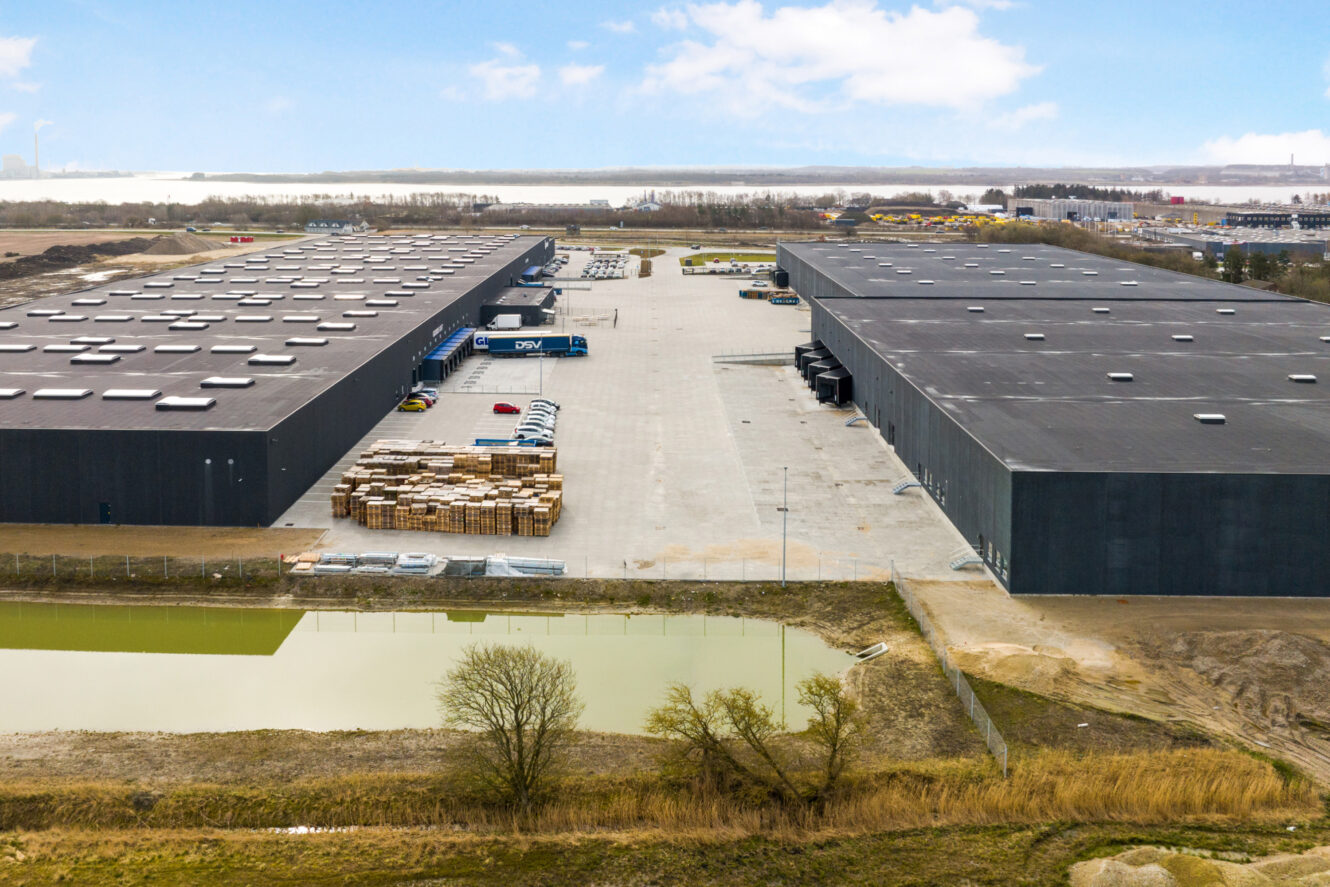 Luftfoto af et stort industrielt lagerkompleks med stablede paller, skibscontainere og parkerede lastbiler, afgrænset af en lille dam og åbent land.