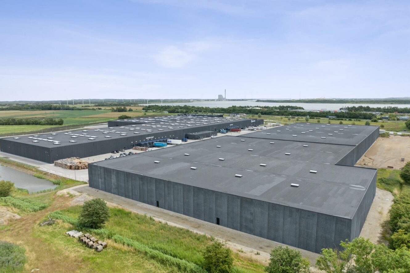 Luftfoto af et stort industrielt lagerkompleks med sorte tage, omgivet af grønne områder, marker og en kanal under en klar himmel.