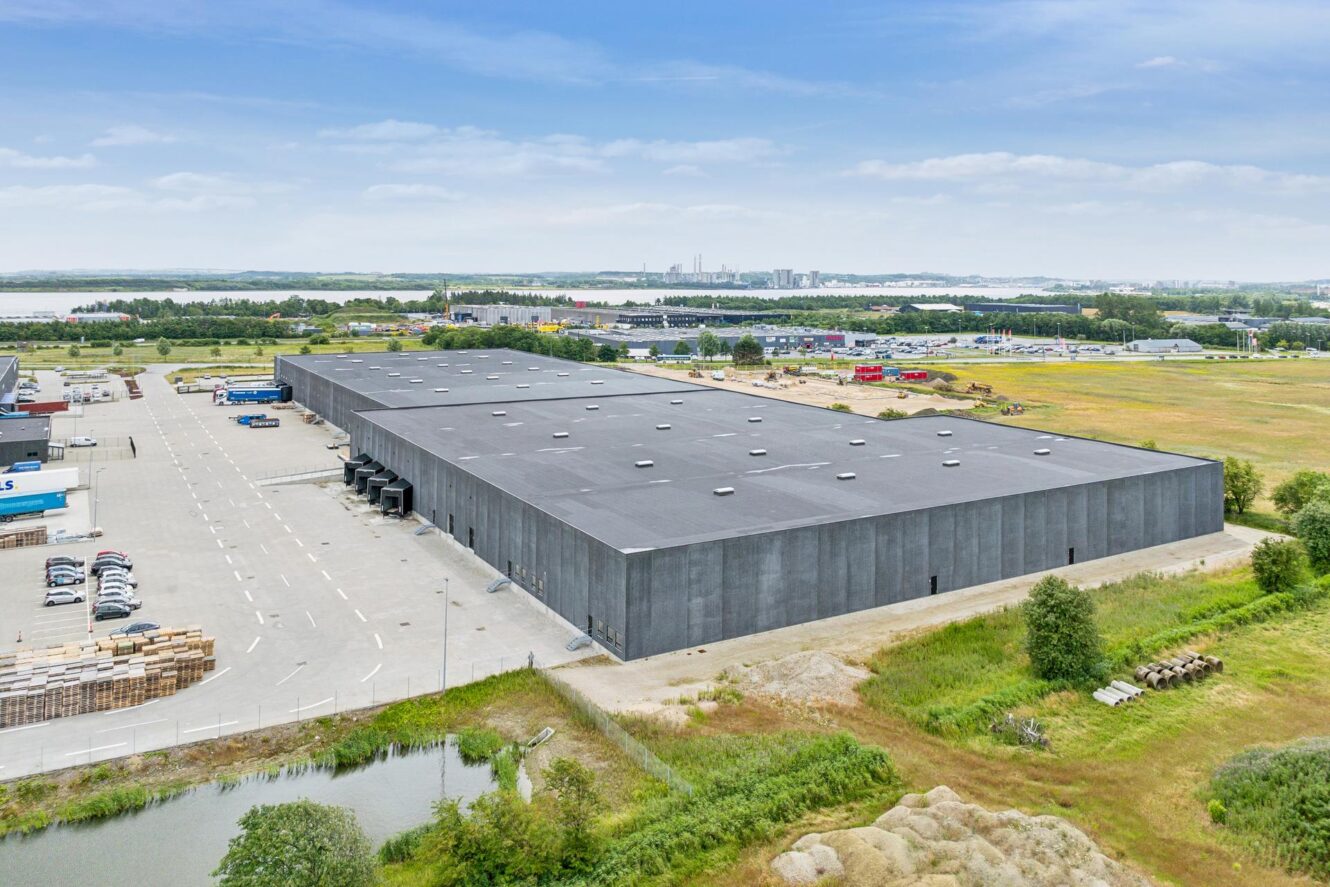 Et stort lager med sort tag er omgivet af parkeringspladser, grønne områder og industribygninger under en delvis overskyet himmel.