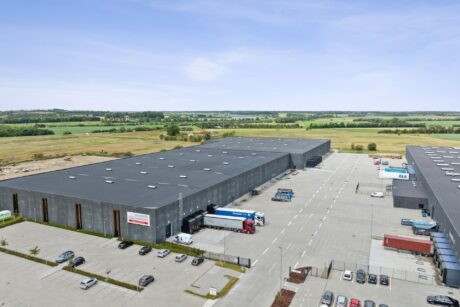 Luftfoto af et stort industrielt lagerkompleks med lasteramper, lastbiler og biler midt i markerne under en delvis overskyet himmel.