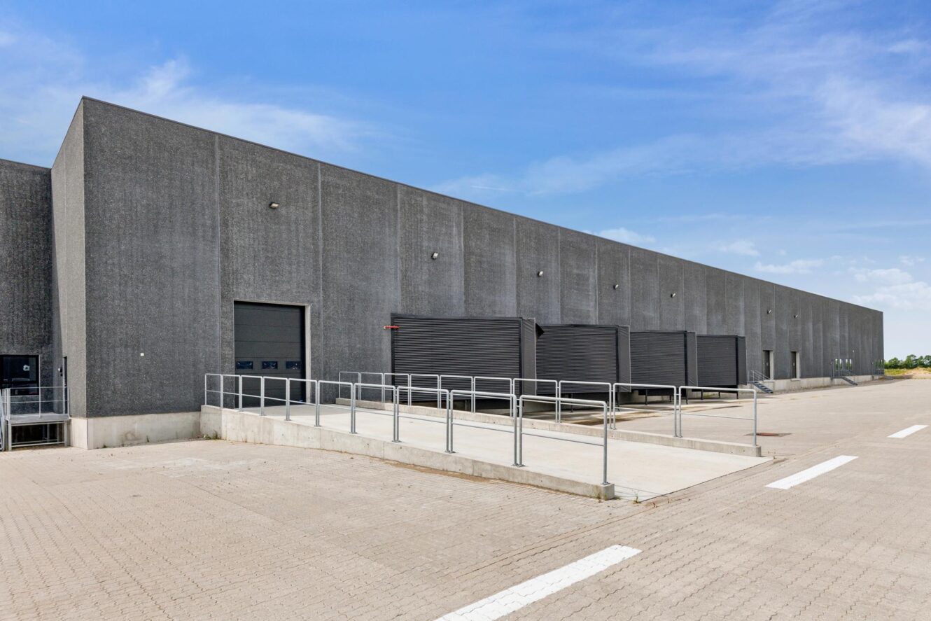 Stort industrilager med flere lasteramper, lukkede døre og en betonrampe under en klar blå himmel.