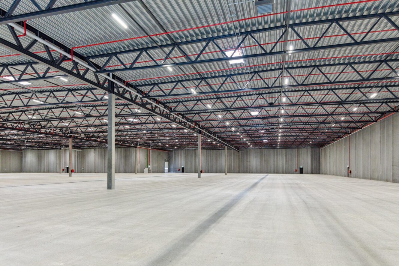 Stort, tomt industrilager med betongulv, højt metal loft, synlige bjælker og grå vægge. Der er hverken udstyr eller opbevarede genstande.