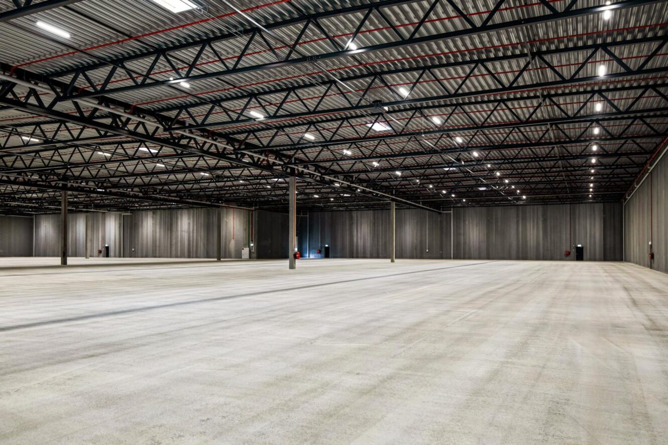 Et stort, tomt lagerlokale med betongulv, højt til loftet, synlige metalbjælker og støttepiller placeret med jævne mellemrum.
