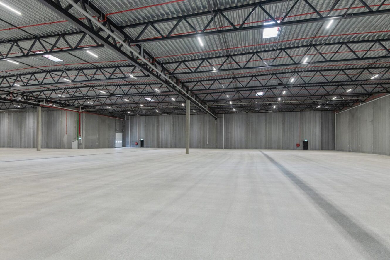 Stort, tomt lagerlokale med betongulv, højt loft af bølgeblik, synlige bjælker og flere udgangsdøre langs væggene.