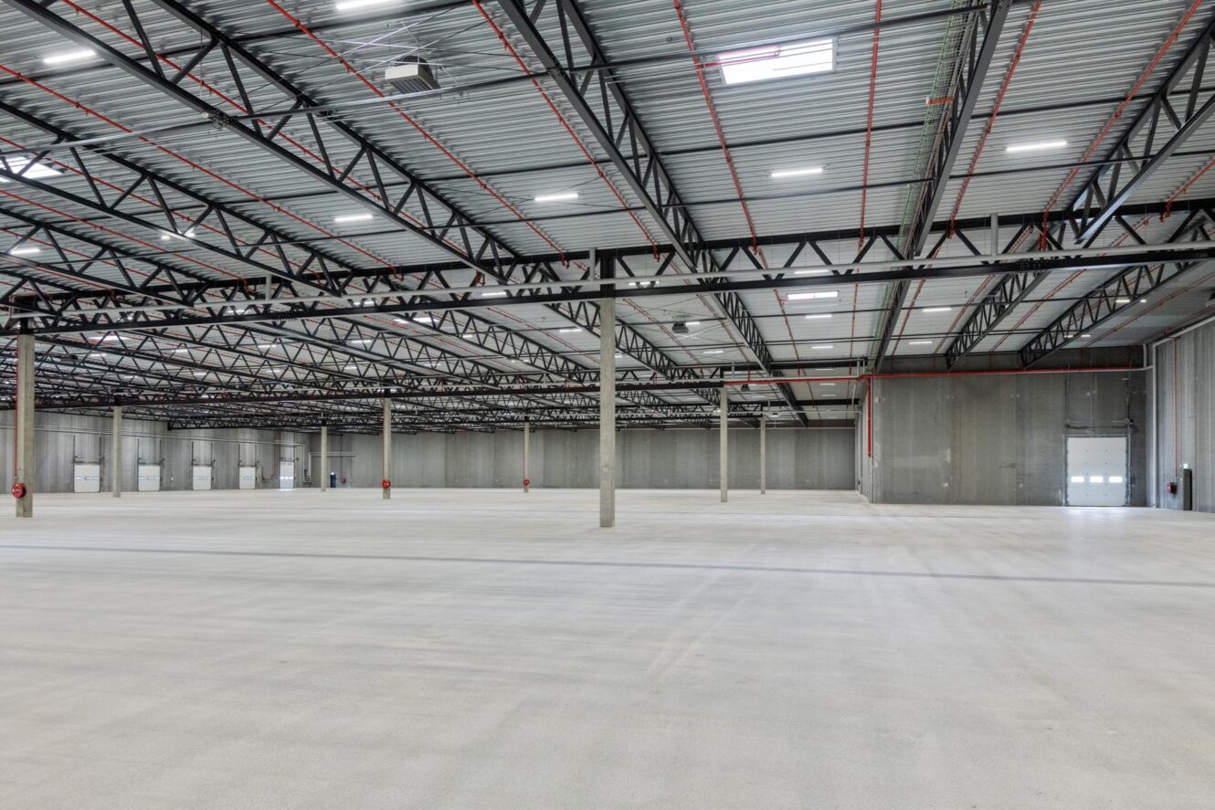 Stort, tomt lager med betongulve, højt metal loft, synlige bjælker og støttepiller. Der er garageporte langs væggene.