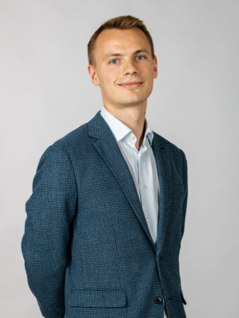 En ung mand i blåternet habitjakke og hvid skjorte står på en almindelig lysegrå baggrund og kigger ind i kameraet med et lille smil.