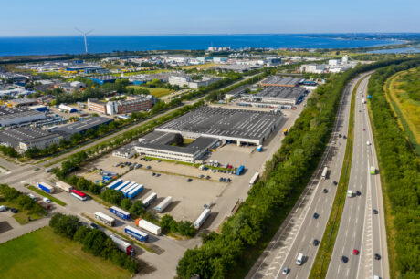 Luftfoto af en industripark med lagerbygninger, lastbiler og parkeringspladser ved siden af en motorvej, med kystlinje, vand og vindmølle i baggrunden.