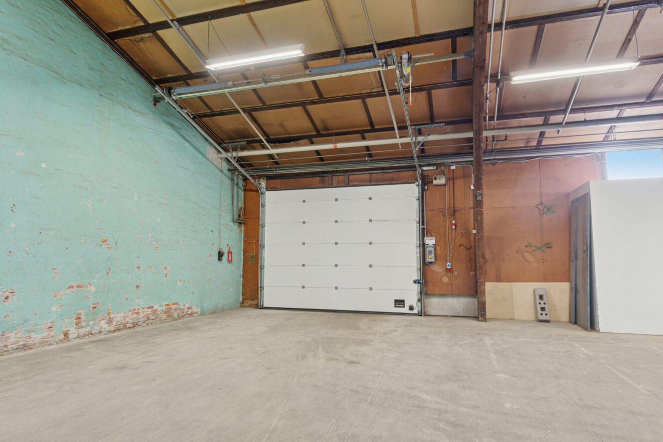 Interiør af et tomt industrilager med en stor garageport, synlige bjælker, mintgrønne og brune vægge og betongulv.