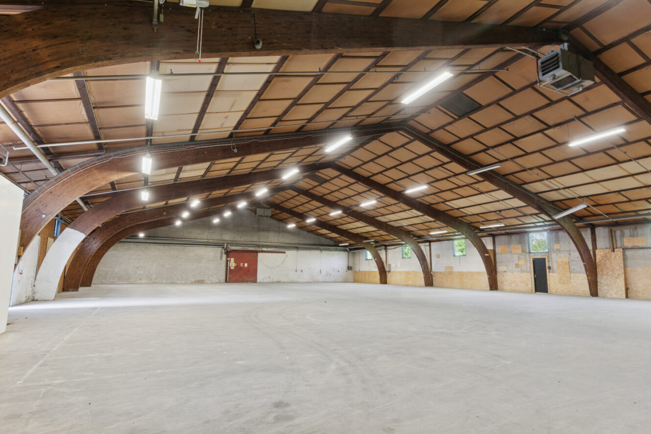 Rummelig, tom lagerbygning med buede træbjælker, synligt loft, betongulv og fluorescerende belysning. Røde dobbeltdøre og vinduer er synlige på den fjerneste væg.