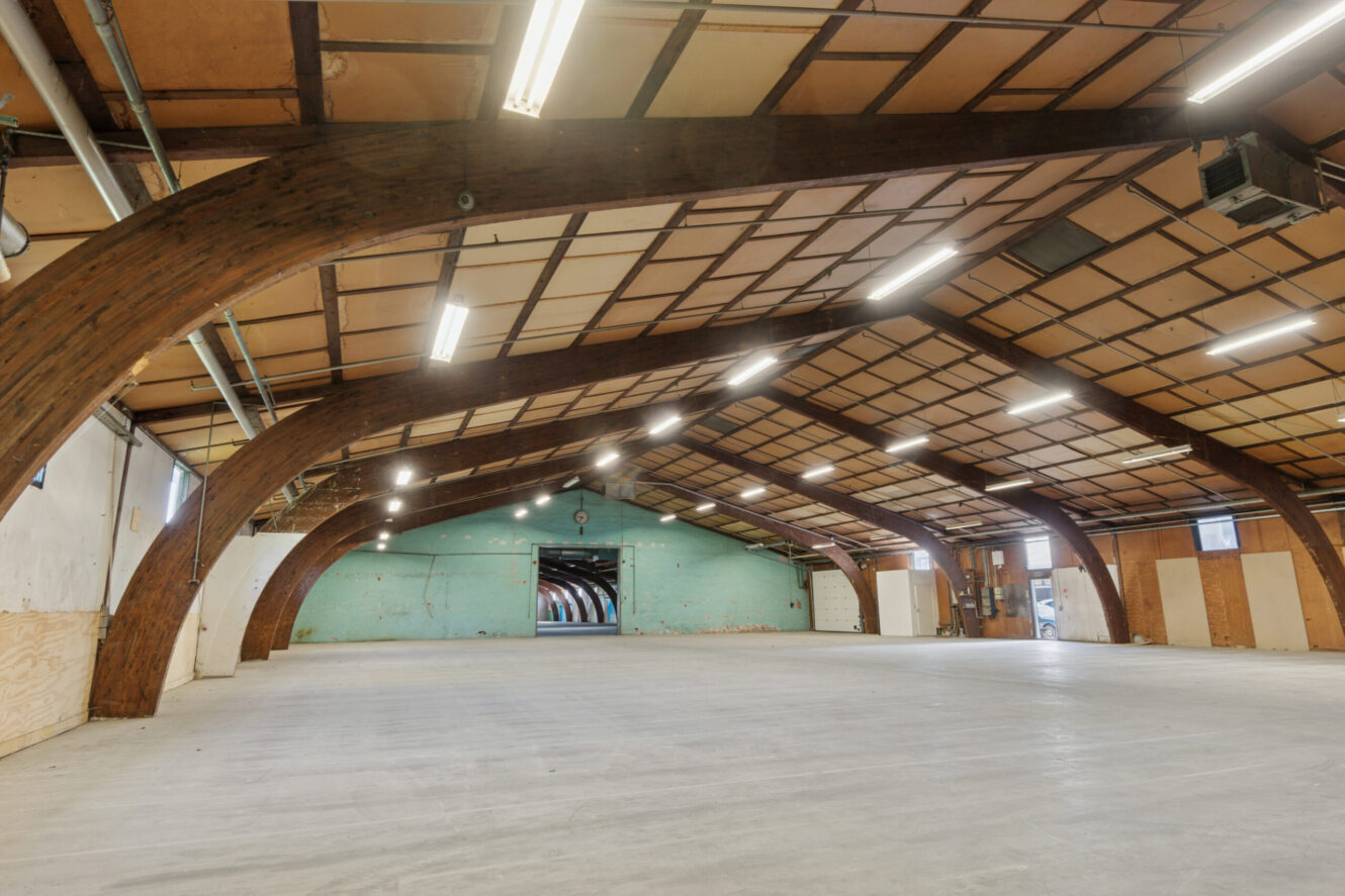 Stor, tom lagerbygning med synlige træbjælker, højt til loftet, betongulv, lysstofrør og en lysegrøn væg i den fjerneste ende.