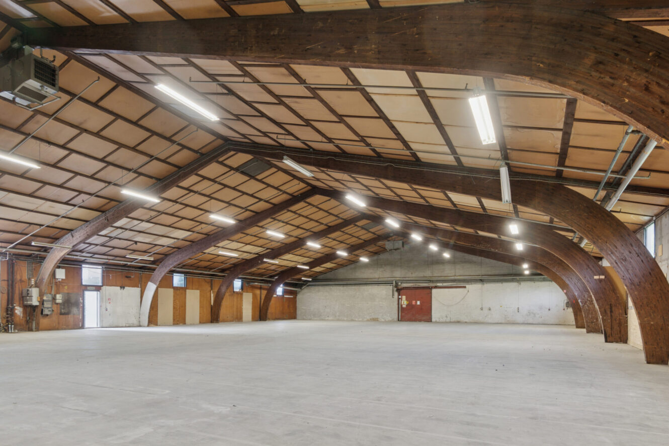 Stor, tom lagerhal med træbjælker, betongulv, hvide vægge og lysstofrør i loftet. Røde dobbeltdøre og forsyningspaneler er synlige langs den ene væg.