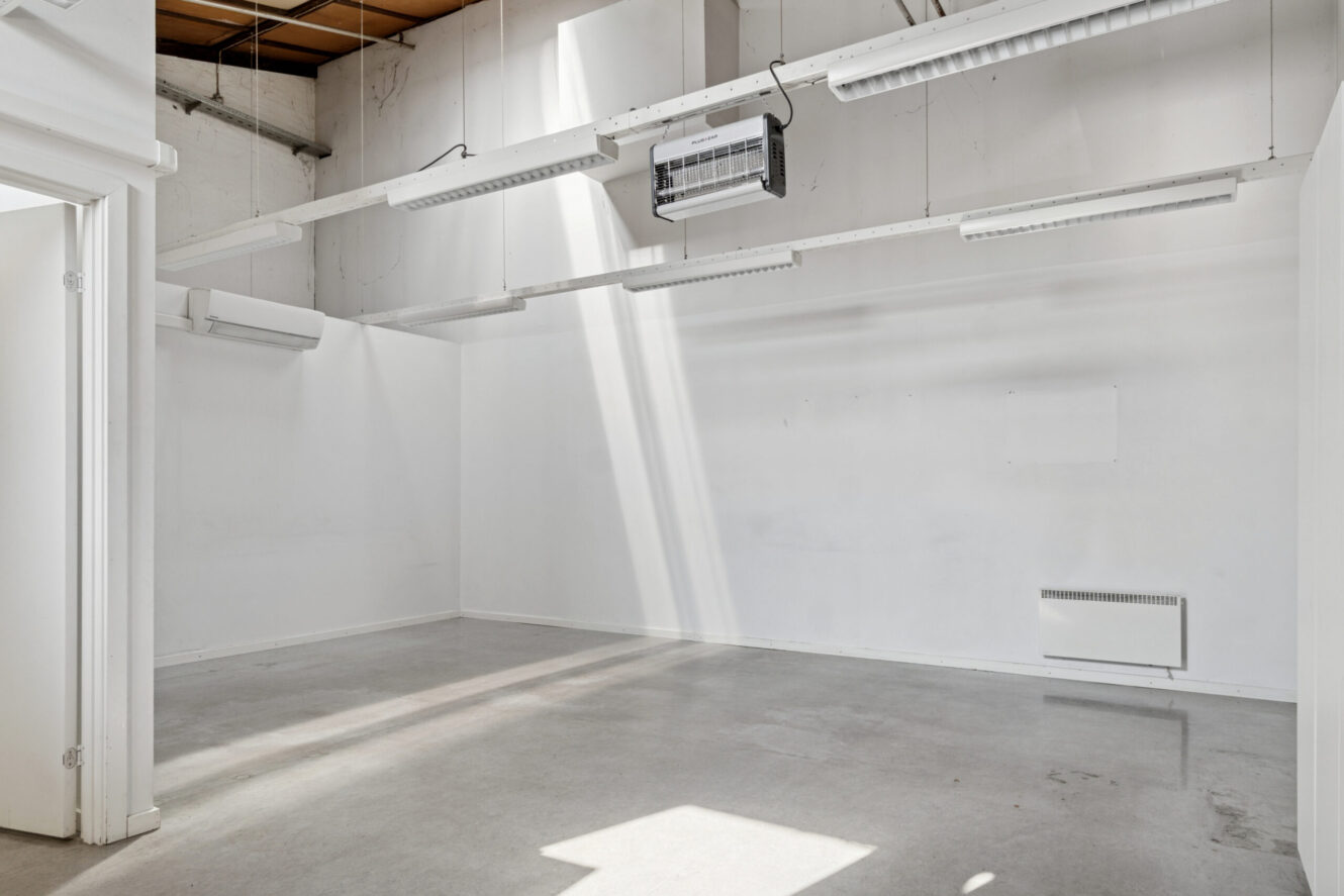 Tomt rum i industriel stil med hvide vægge, betongulv, synligt loft og lysstofrør. Sollyset strømmer ind gennem et højt vindue.