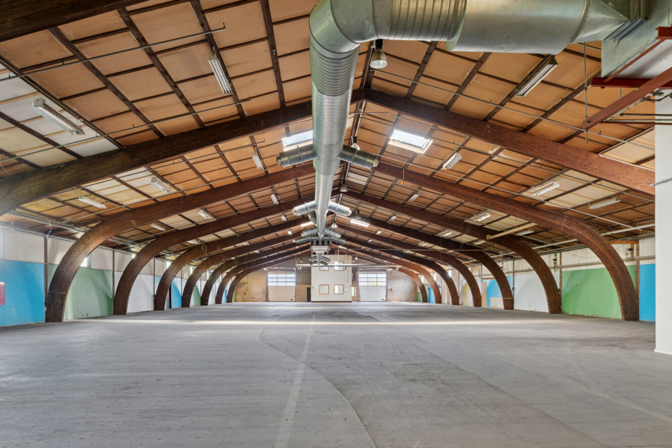 Stort, tomt industrilager med buede træbjælker, synlige kanaler, betongulv og turkis- og hvidmalede vægge. Naturligt lys kommer ind gennem vinduer i den fjerneste ende.