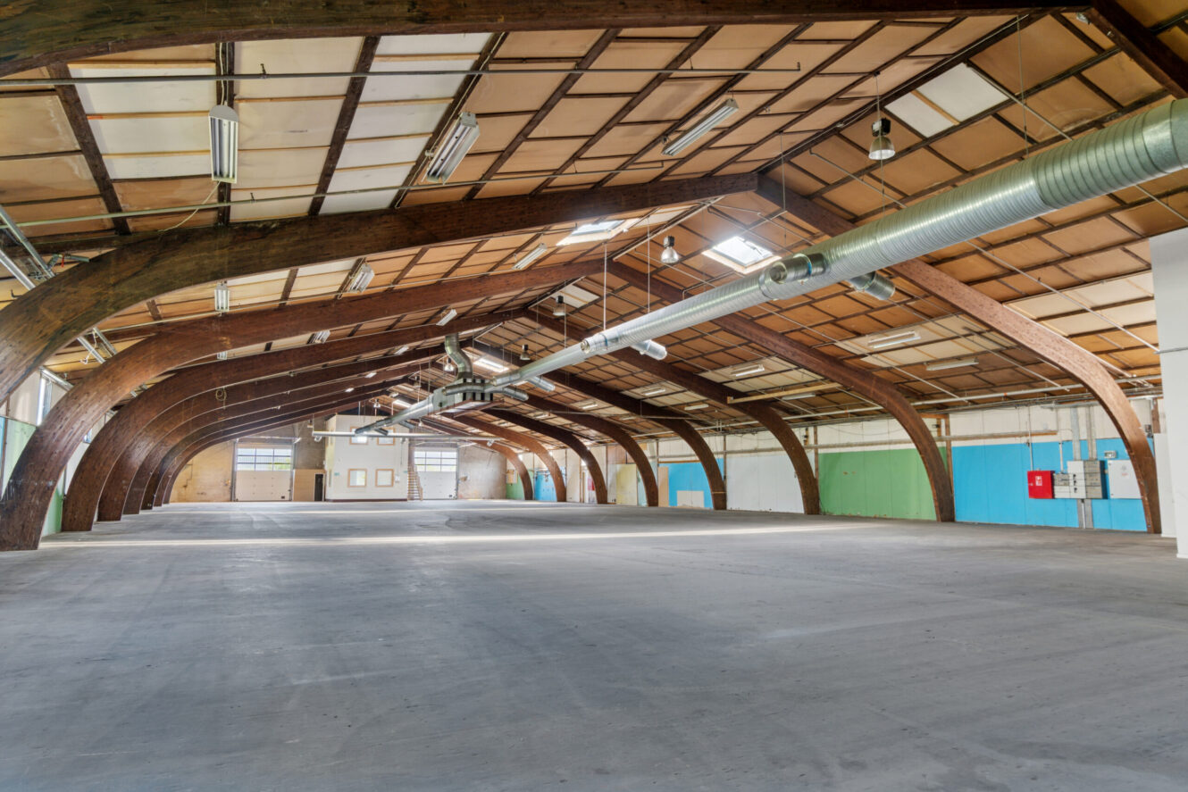 Rummelig, tom lagerbygning med buede træbjælker, synlige kanaler og betongulv med store vinduer, ovenlys og blågrønne og hvide vægge.