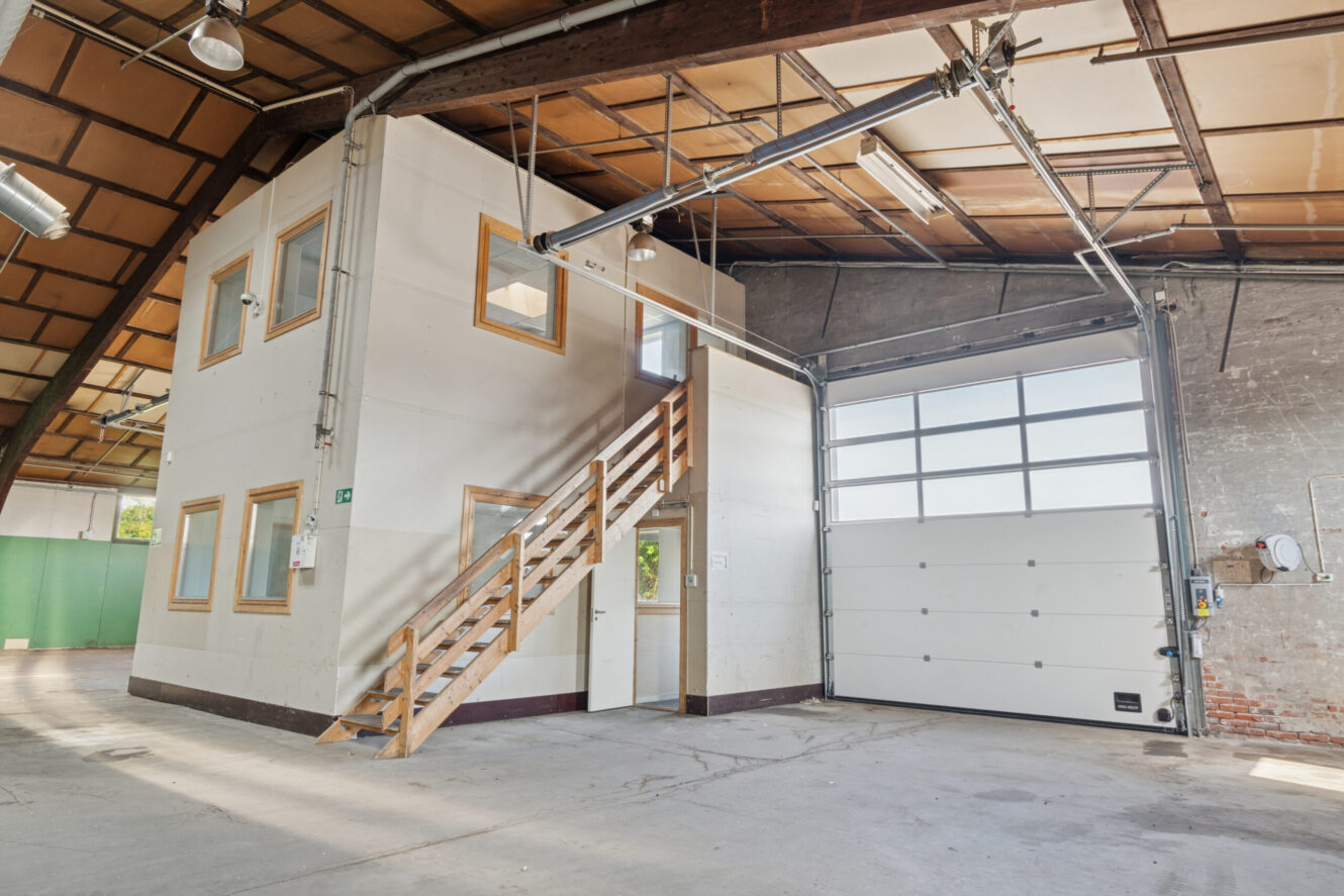 Interiør af et industrilager med en lille kontorbygning i to etager, trætrapper, stor garageport og synlige bjælker.