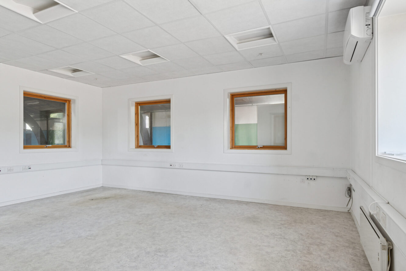 Tomt, umøbleret rum med hvide vægge, tre træindrammede vinduer, et gråt gulv, lysstofrør i loftet og en vægmonteret airconditionenhed.