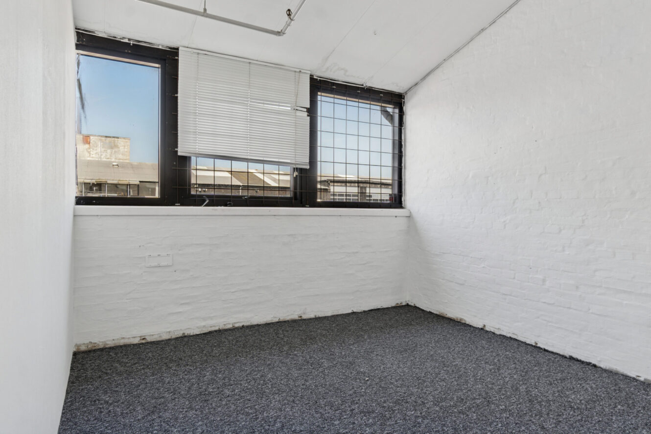 Lille tomt rum med hvidmalede murstensvægge, to store vinduer med persienner og metalstænger og et gråt gulvtæppe.