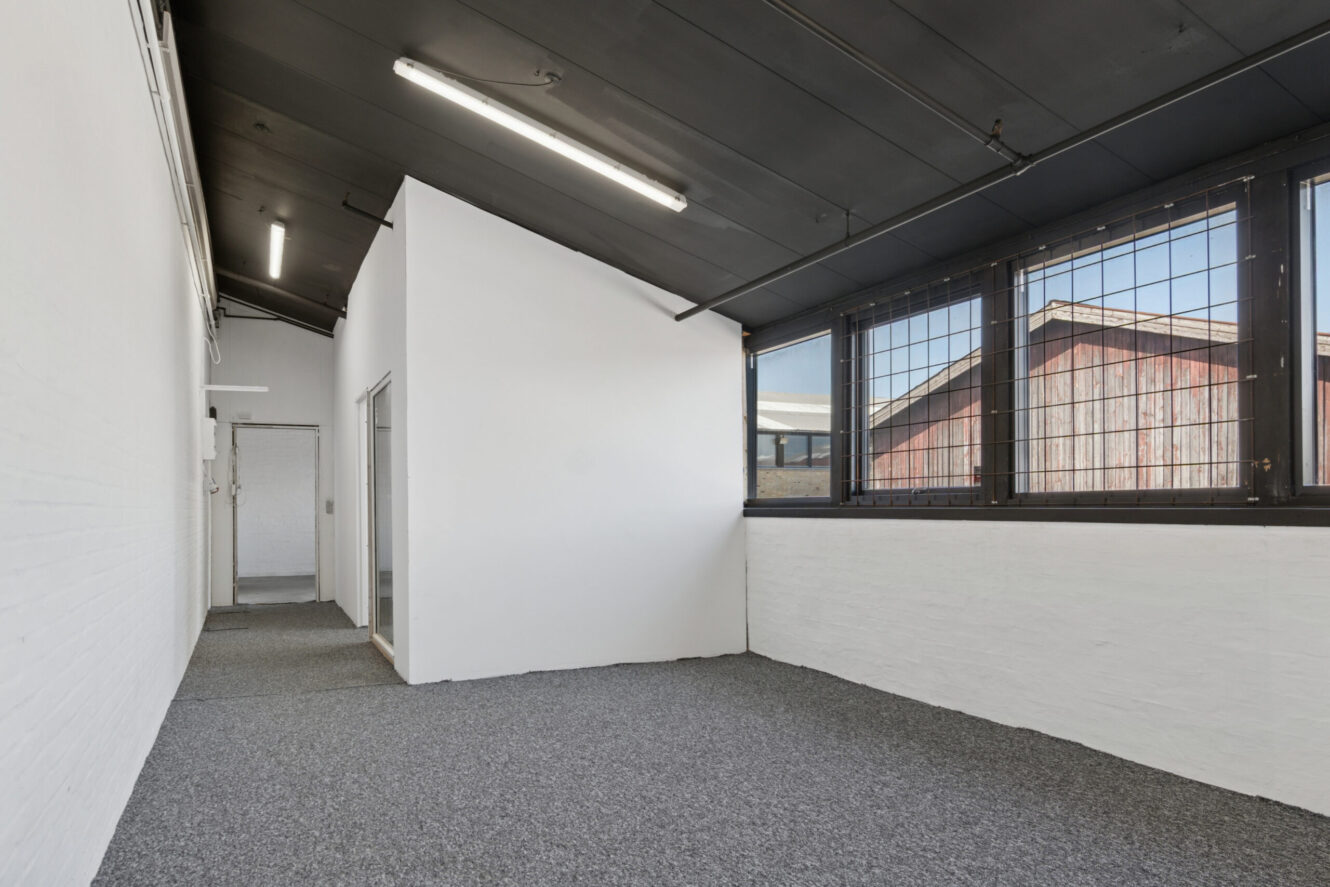 Umøbleret værelse med hvide vægge, mørkt loft, gråt gulvtæppe og store vinduer med metalstænger med udsigt til en nabobygning.