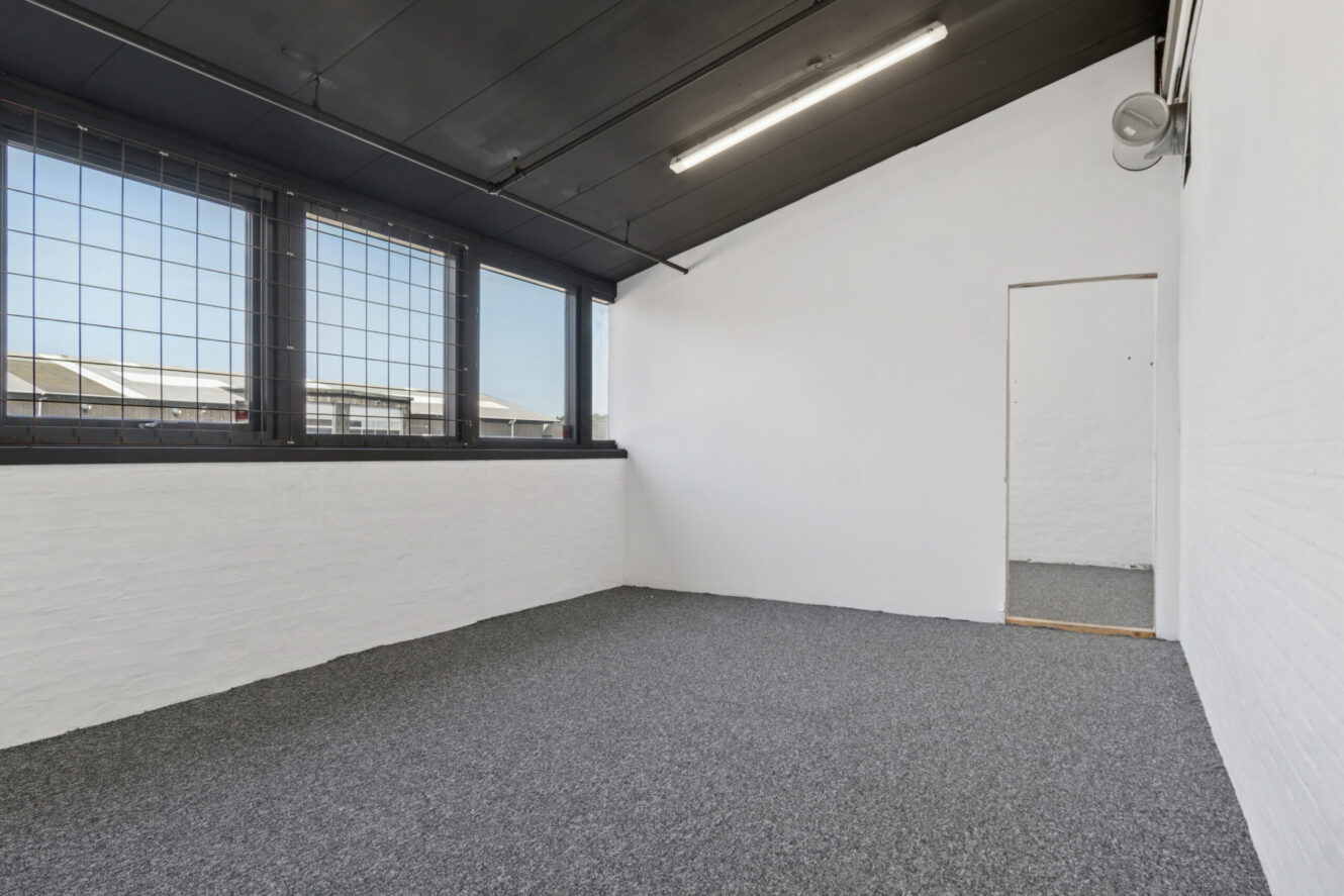 Umøbleret rum med hvide vægge, gråt gulvtæppe, store vinduer med sikkerhedsgitre og en enkelt åben dør.