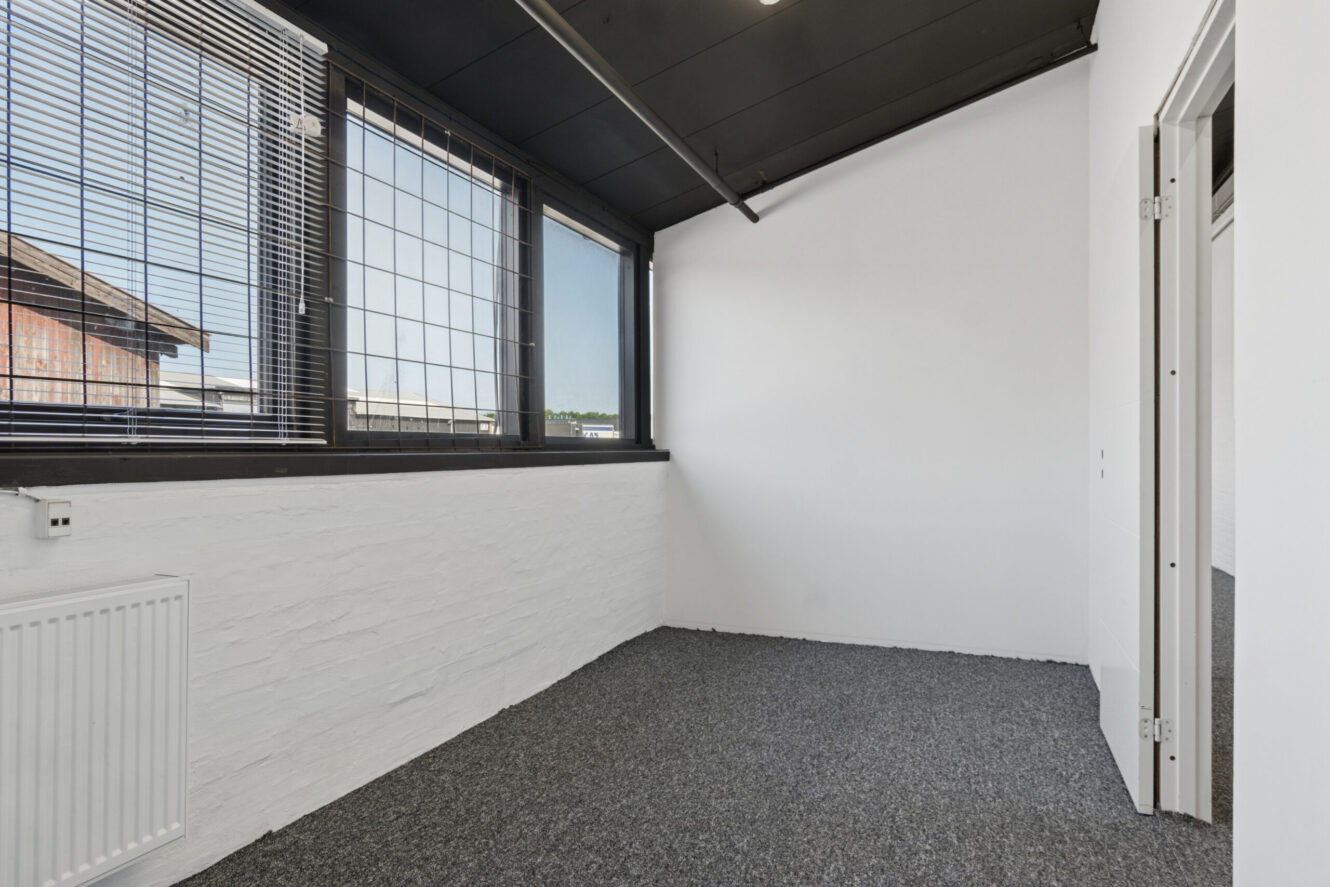 Lille, tomt rum med hvide vægge, gulvtæppe, store vinduer med persienner og tremmer og en delvis åben hvid dør.