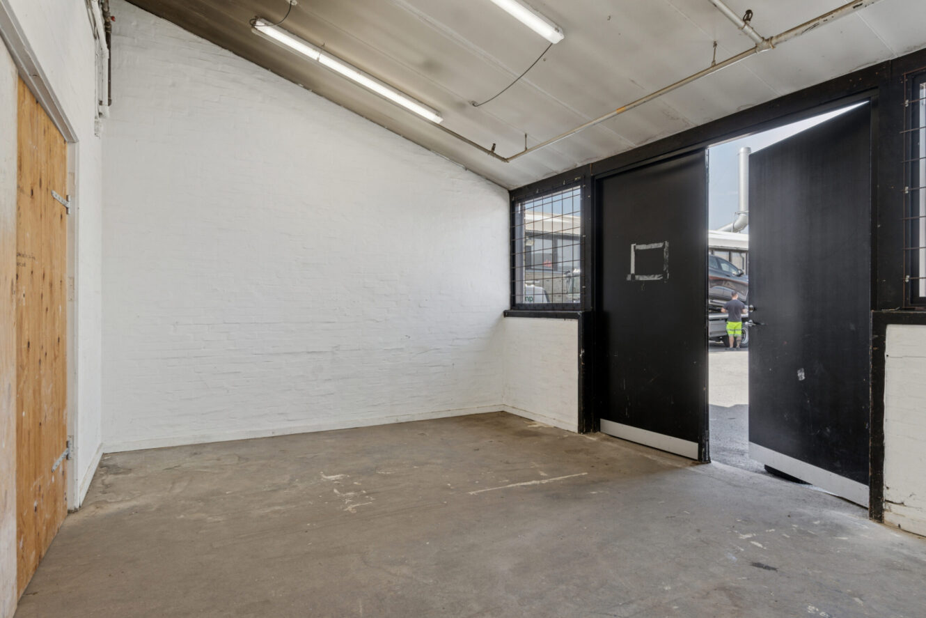 Tomt industrilokale med hvide murstensvægge, betongulv, lysstofrør, en trædør til venstre og sorte dobbeltdøre, der åbner ud til et udendørs område.
