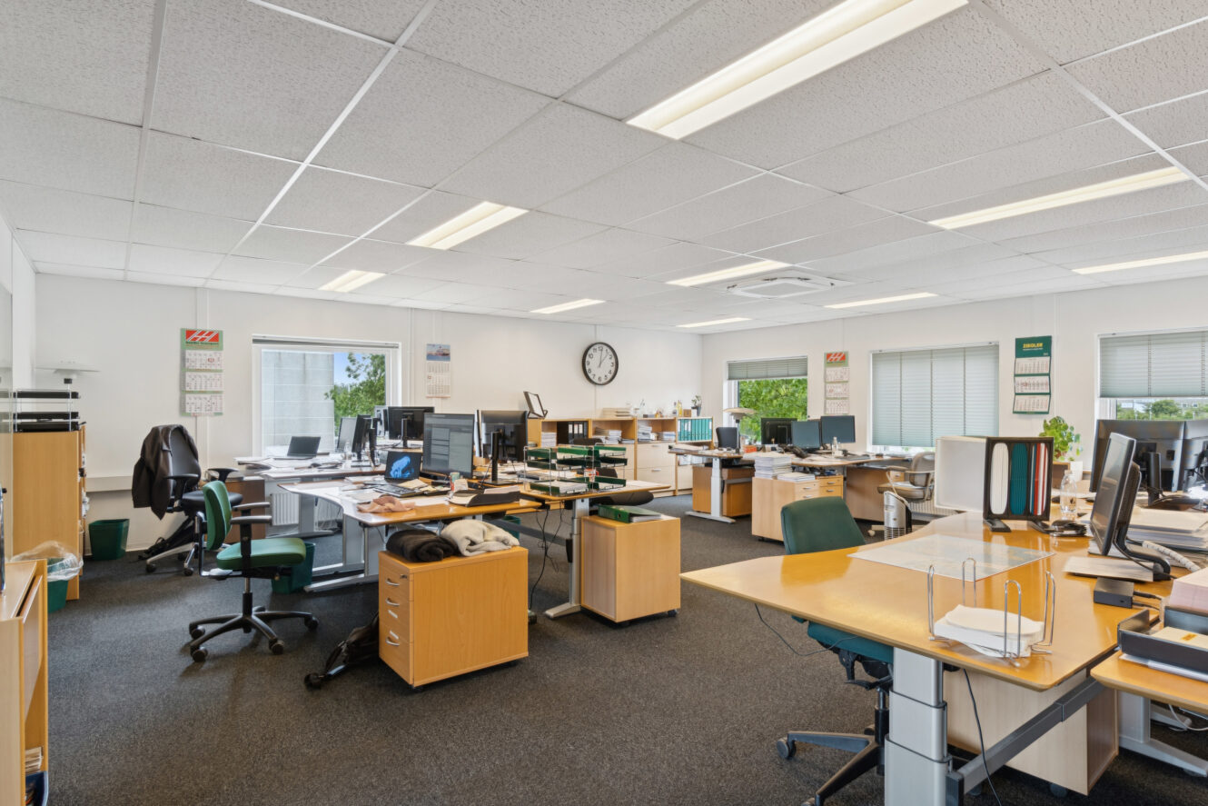 Åbent kontor med flere skriveborde, computere, kontorstole, papirer og materialer, oplyst af loftslamper og naturligt lys fra vinduer.