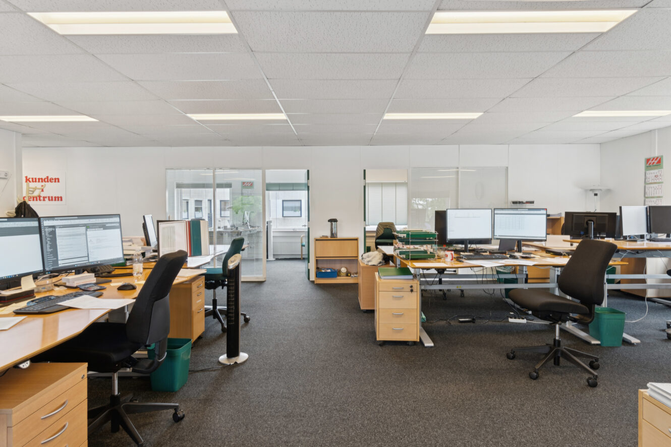 Moderne åbent kontorlandskab med skriveborde, stole, computere og kontorartikler. Store vinduer og glasskillevægge giver naturligt lys. Rummet er ryddeligt og organiseret.