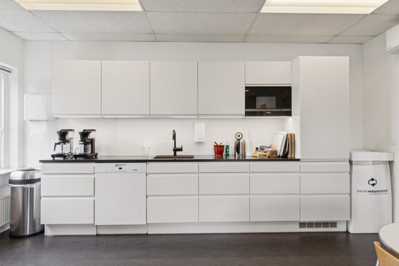 Moderne kontorkøkken med hvide skabe, sort bordplade, kaffemaskiner, en vask, en mikroovn og en genbrugsspand. Rummet er rent og velorganiseret.