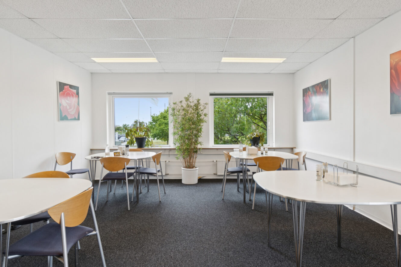 Et lyst, rent pauserum med flere borde og stole, en potteplante mellem to store vinduer og blomsterkunst på de hvide vægge.