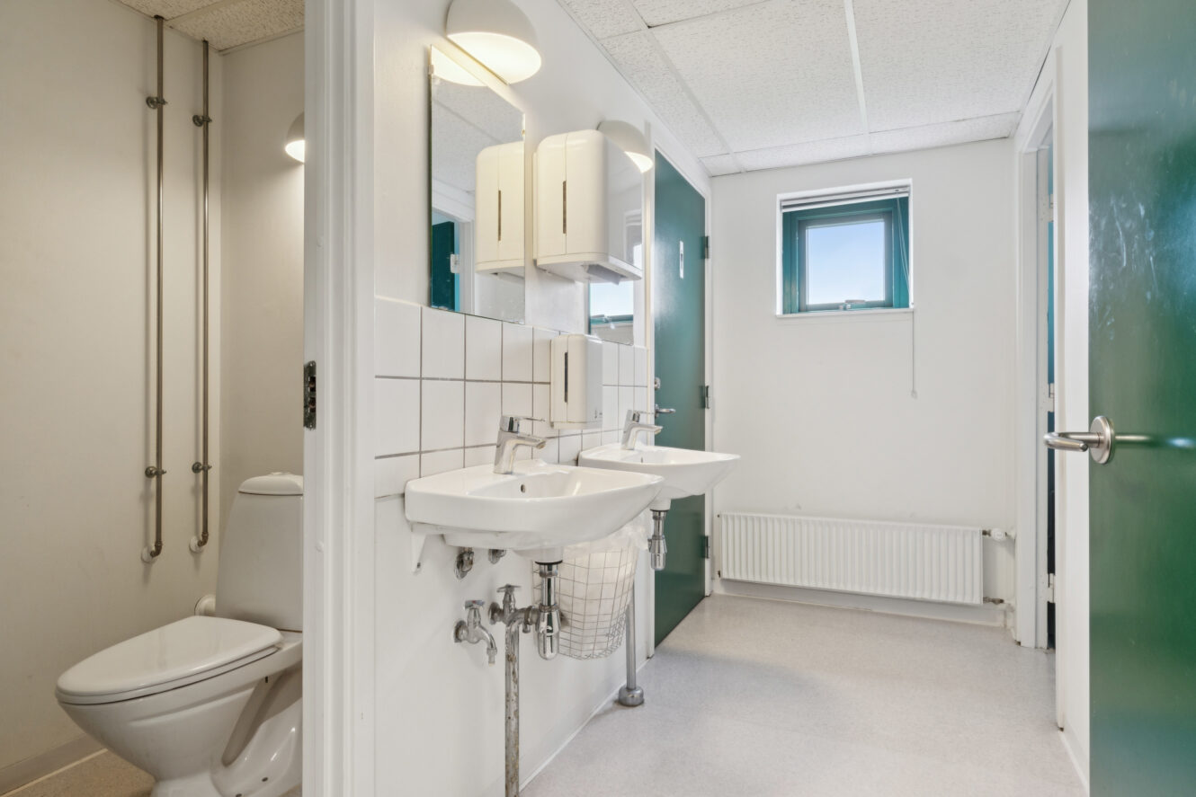 Et rent, moderne badeværelse med to håndvaske, et væghængt spejl, et toilet, et vindue og grønne døre. Hvide fliser og neutrale farver dominerer rummet.