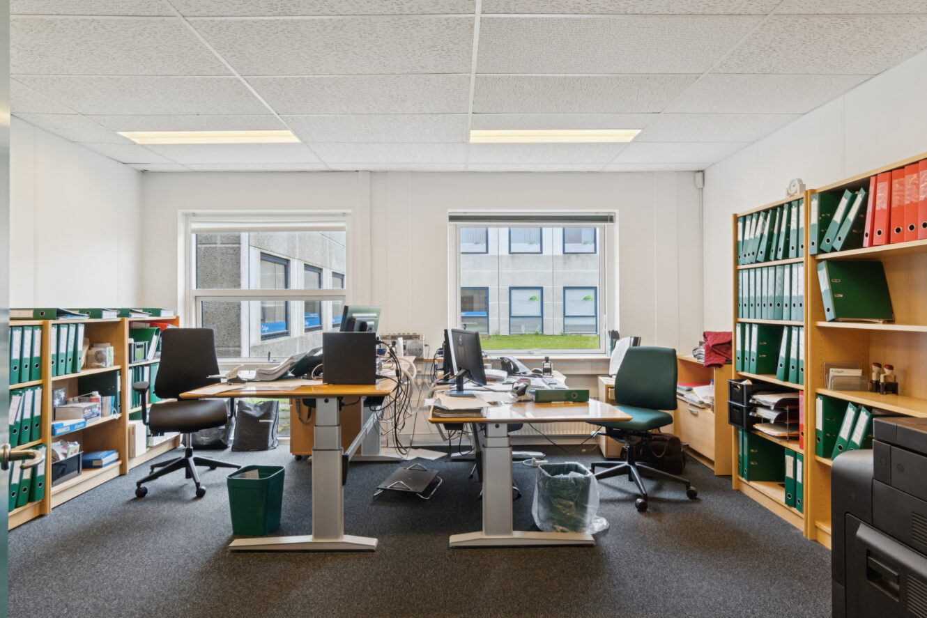 Et moderne kontor med to skriveborde, kontorstole, computere, bogreoler med mapper og store vinduer, der lukker naturligt lys ind.