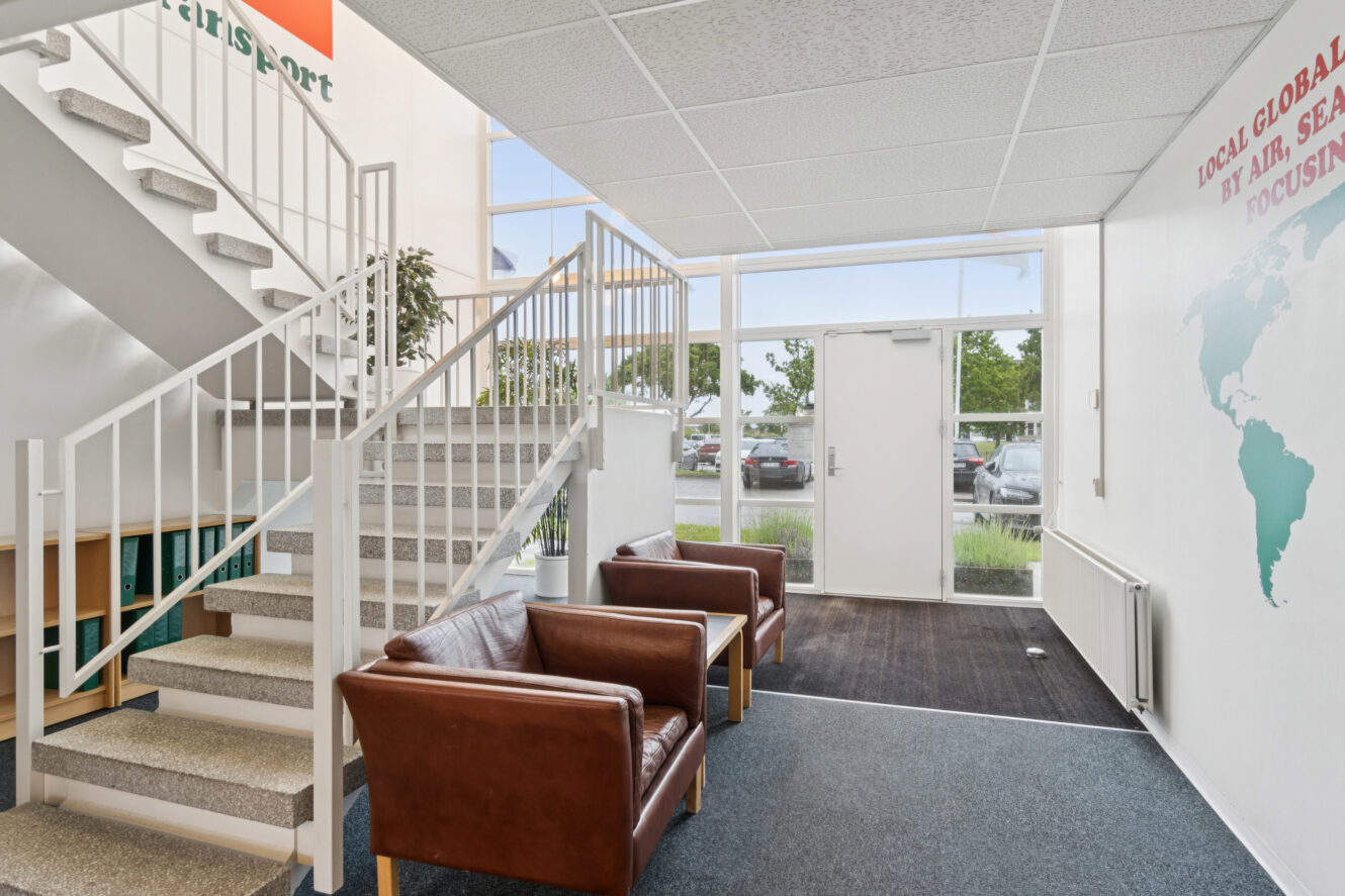 En lille kontorlobby med to brune læderstole, en trappe til venstre, store vinduer og en glasdør, der fører ud.