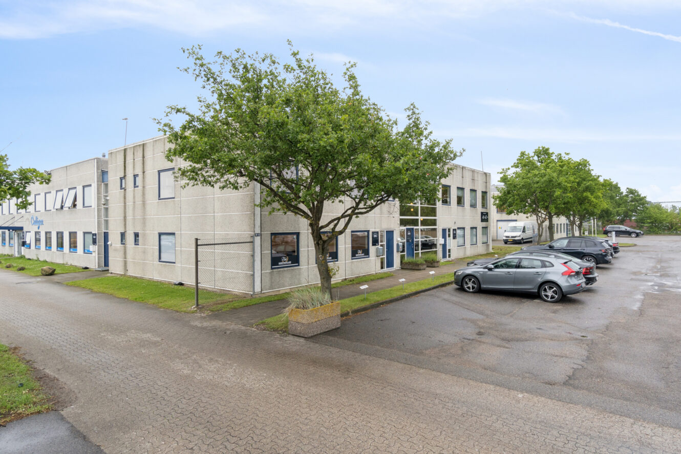 Toetagers kontorbygning med store vinduer, omgivet af træer og parkerede biler på en asfalteret plads under en delvis overskyet himmel.