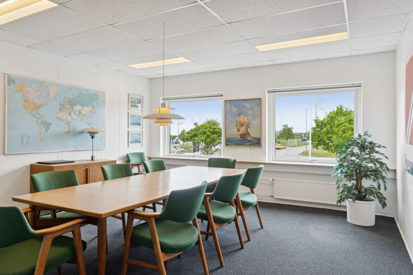 Et konferencelokale med træbord, grønne stole, vægkunst, store vinduer med udsigt til det fri og en potteplante i hjørnet.