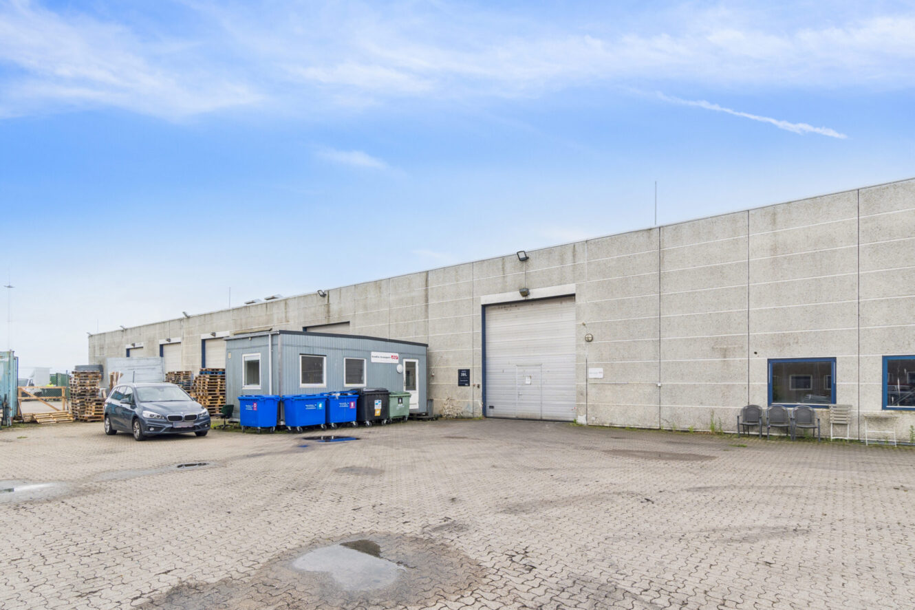 Et lagerhusfacade med en stor garageport, en lille kontortrailer, blå containere, stablede paller og en parkeret bil på en asfalteret grund under en delvist overskyet himmel.