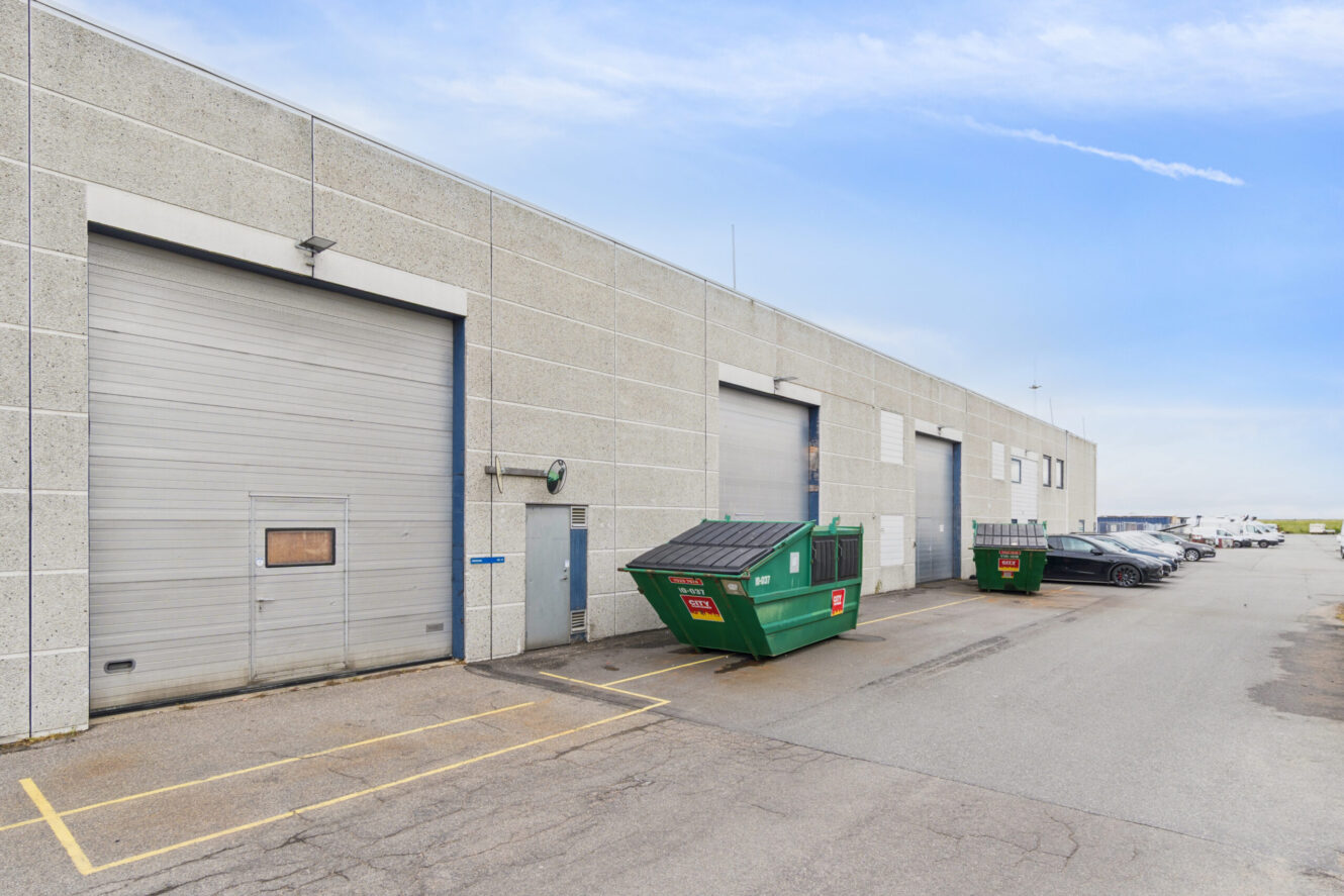 Udvendig udsigt over et lager med lukkede garagedøre, grønne containere, parkerede biler og en klar himmel over.