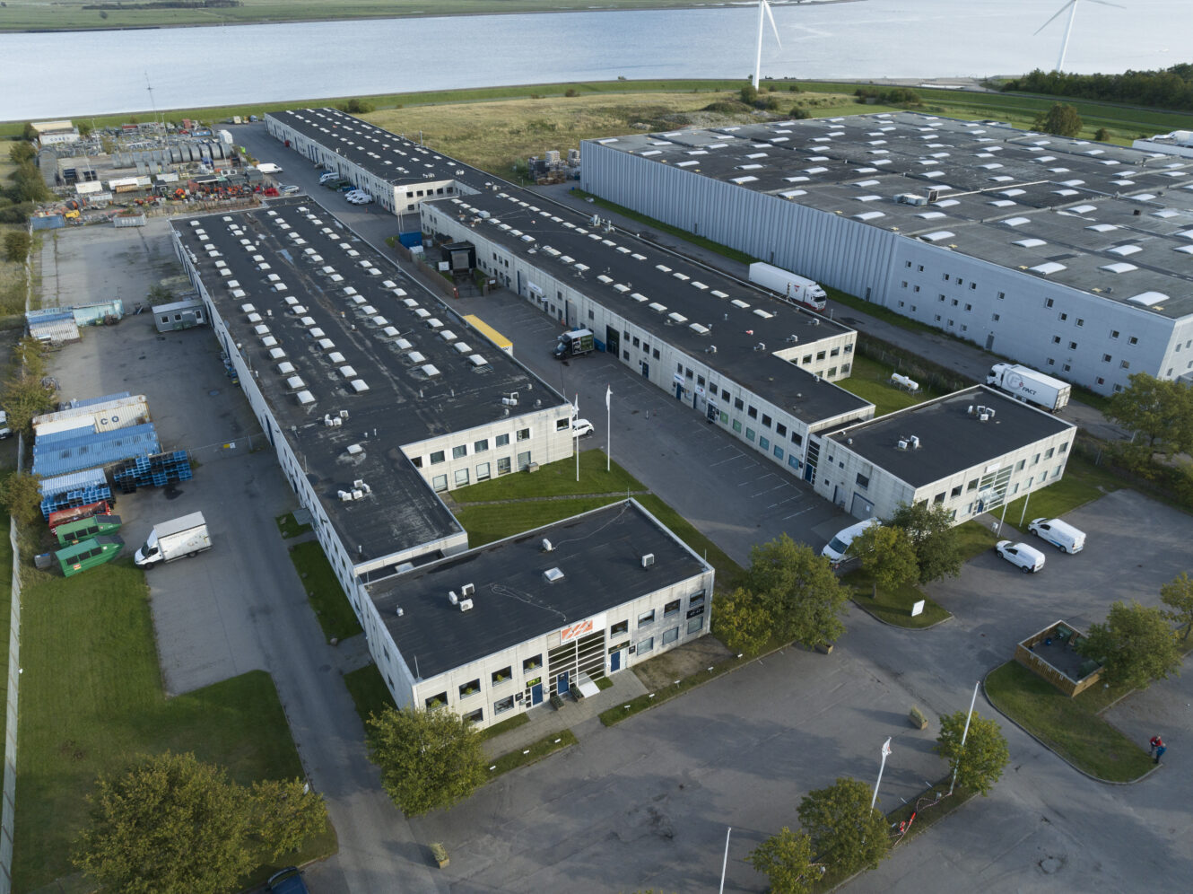 Luftfoto af et industrikompleks med flere store lagerbygninger, tilstødende parkeringsområder, lastbiler og udstyr, der ligger tæt på et vandområde og vindmøller.