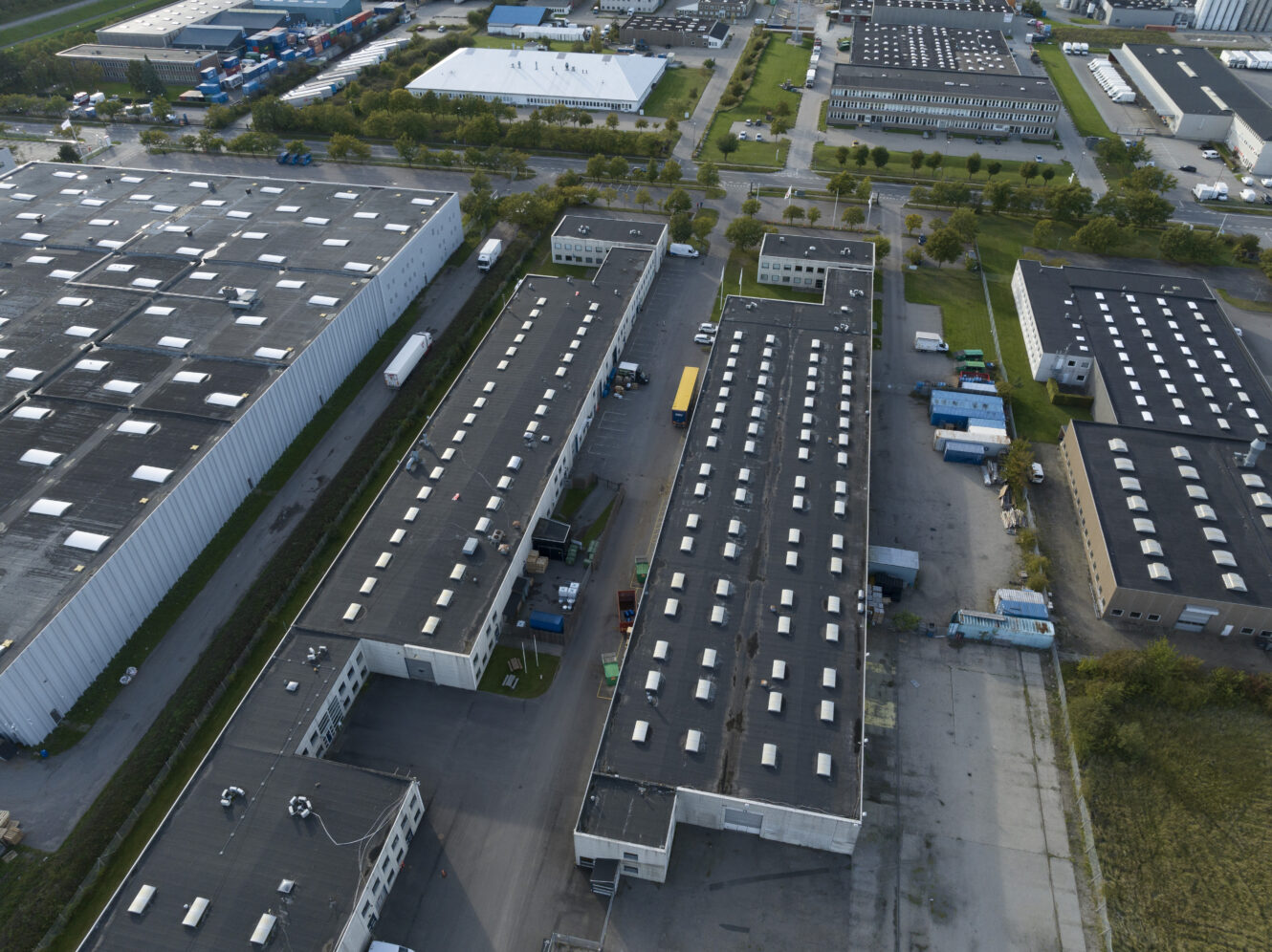 Luftfoto af et industrikompleks med flere store lagerbygninger, parkerede lastbiler og tilstødende parkeringspladser omgivet af veje og grønne områder.