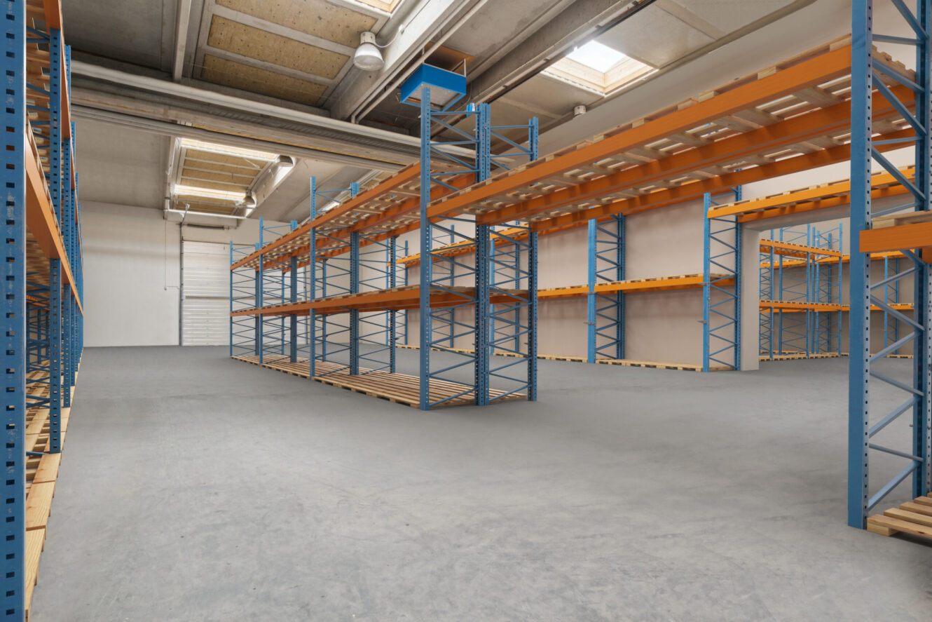 Tomt lagerinteriør med metalreoler, betongulv, højt til loftet og fluorescerende belysning.